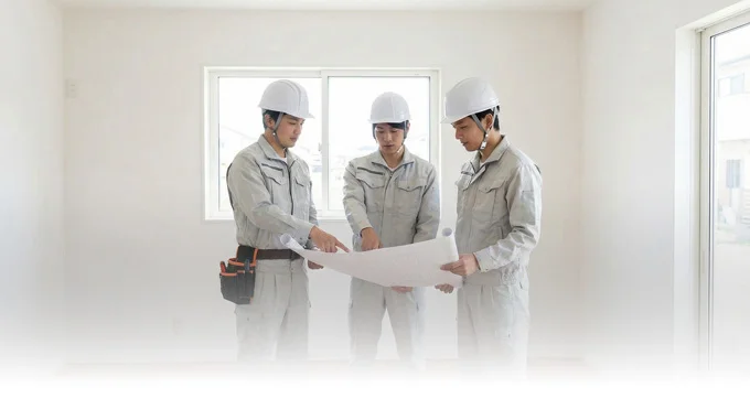 完成間近の部屋で、3人の電気工事士が図面を見ながら打ち合わせをしている様子です。計画性とコミュニケーションの重要性を伝えます。